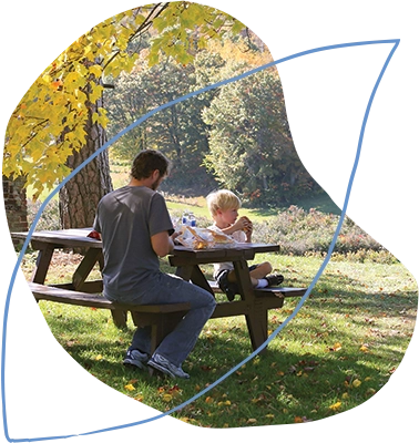 organic shape cut-out photo of a man and his son eating lunch at a picnic table in the fall; the image is overlaid by a light blue line drawing shaped like a leaf
